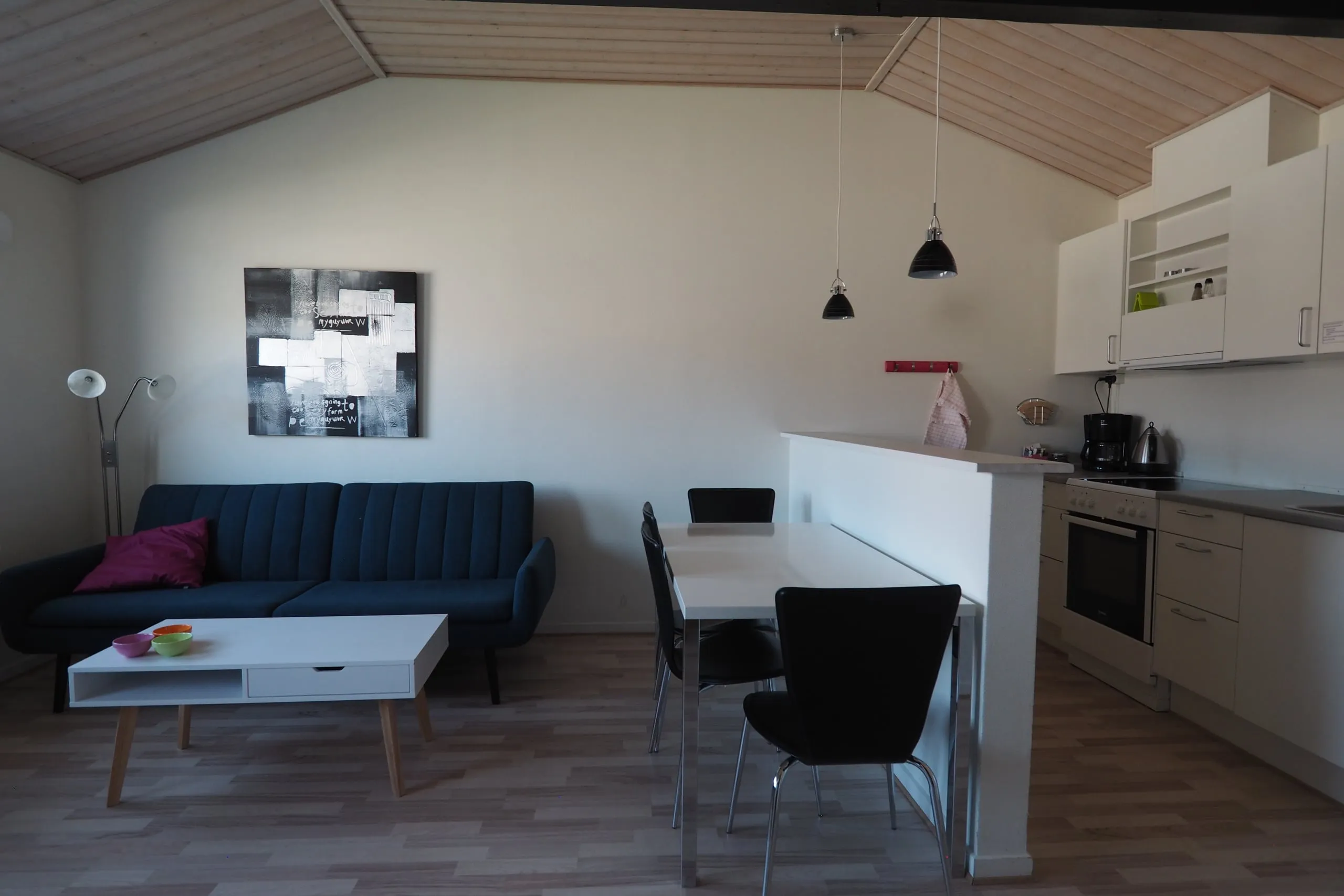 An apartment living room: a blue sofa and coffee table, a dining table, and the kitchen behind a low counter.