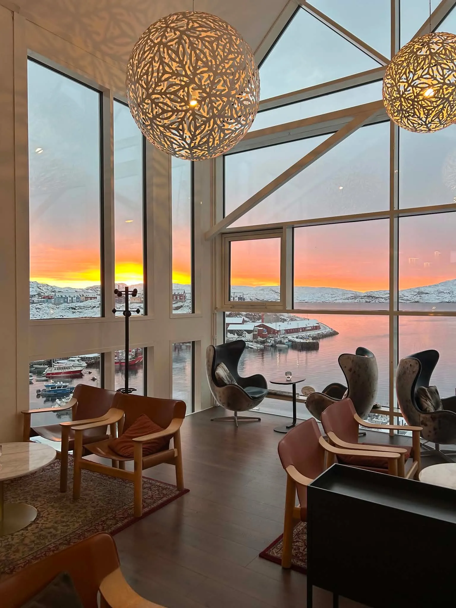 The lounge at sunset, armchairs under patterned pendant lamps before tall windows over the snowy harbour.