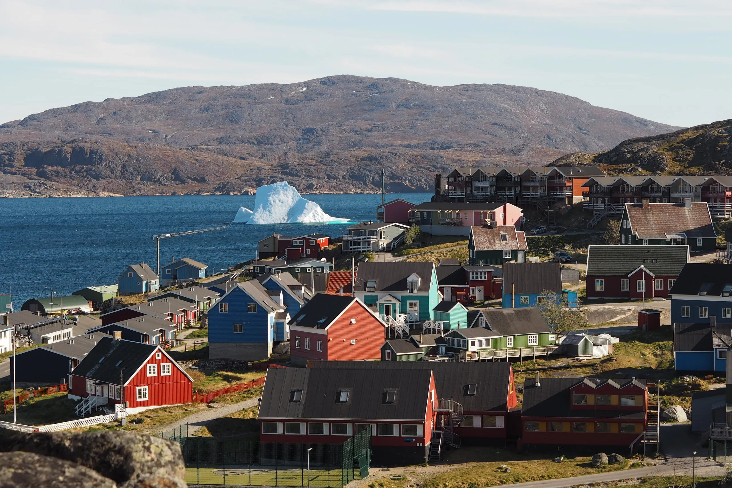 The coloured houses of Qaqortoq running down to the fjord, with an iceberg in the water and hills beyond.