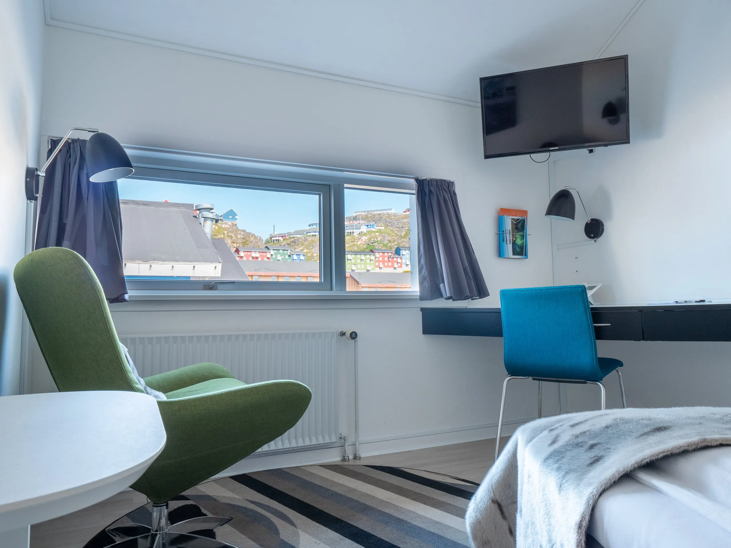 A single room with a green armchair, a blue desk chair and a TV, and a window over the coloured houses of the town.