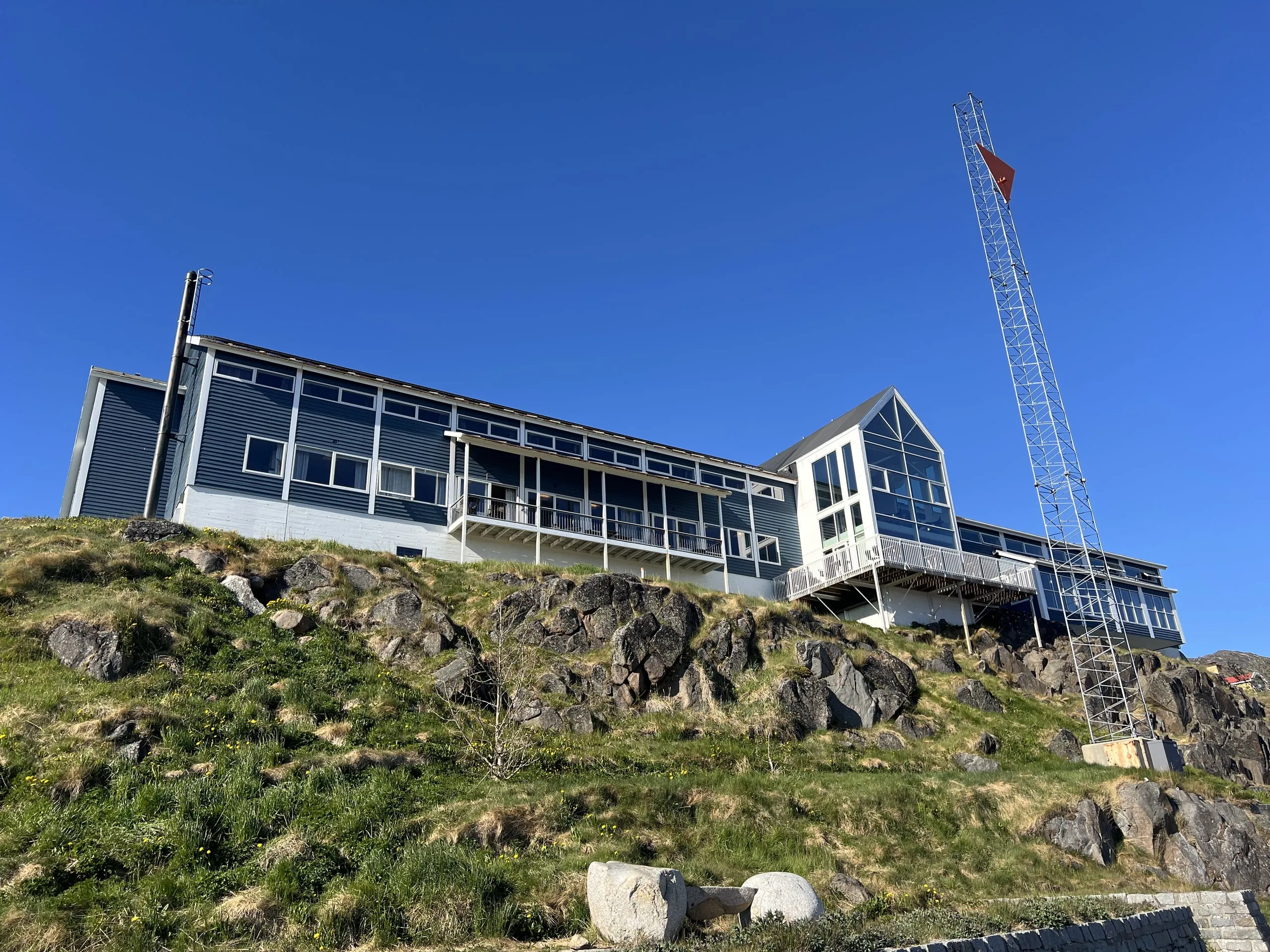 The blue hotel and its tall glass gable on a grassy rock, under a clear blue sky.