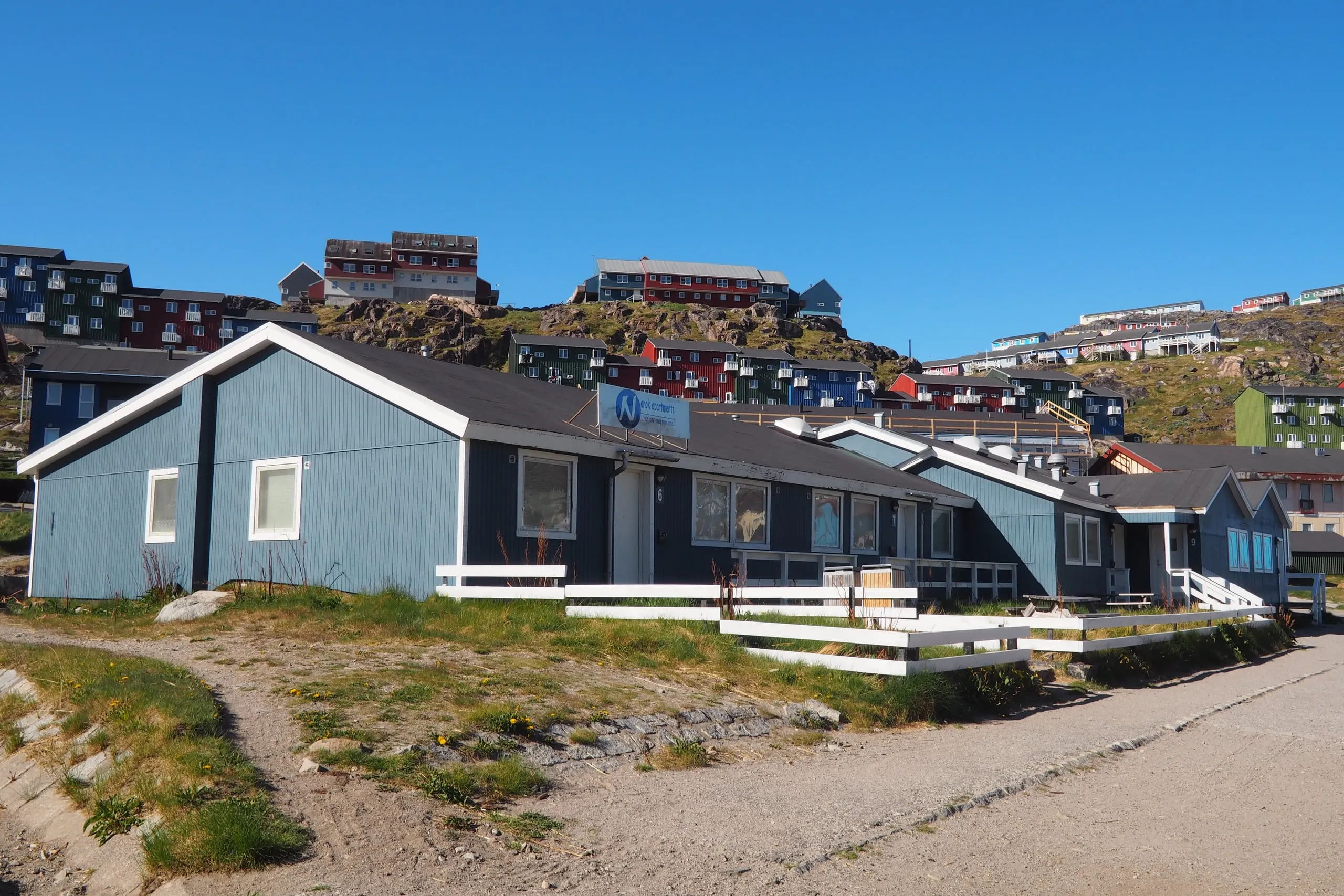 The apartment building, a long blue-grey house with white fences, below the coloured houses of Qaqortoq.
