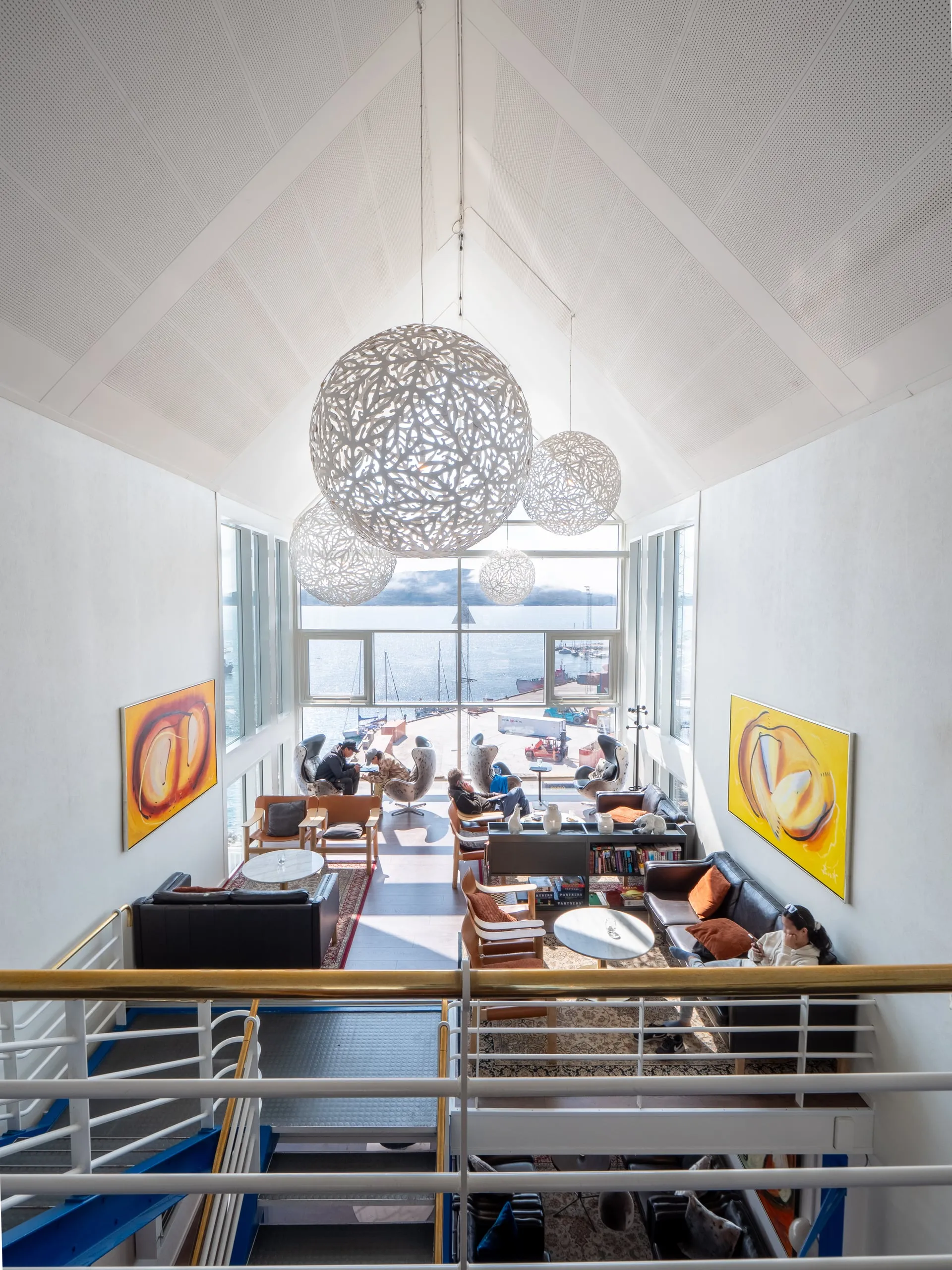 The lounge seen from the gallery above, with round pendant lamps, bright paintings and sofas by a tall window.
