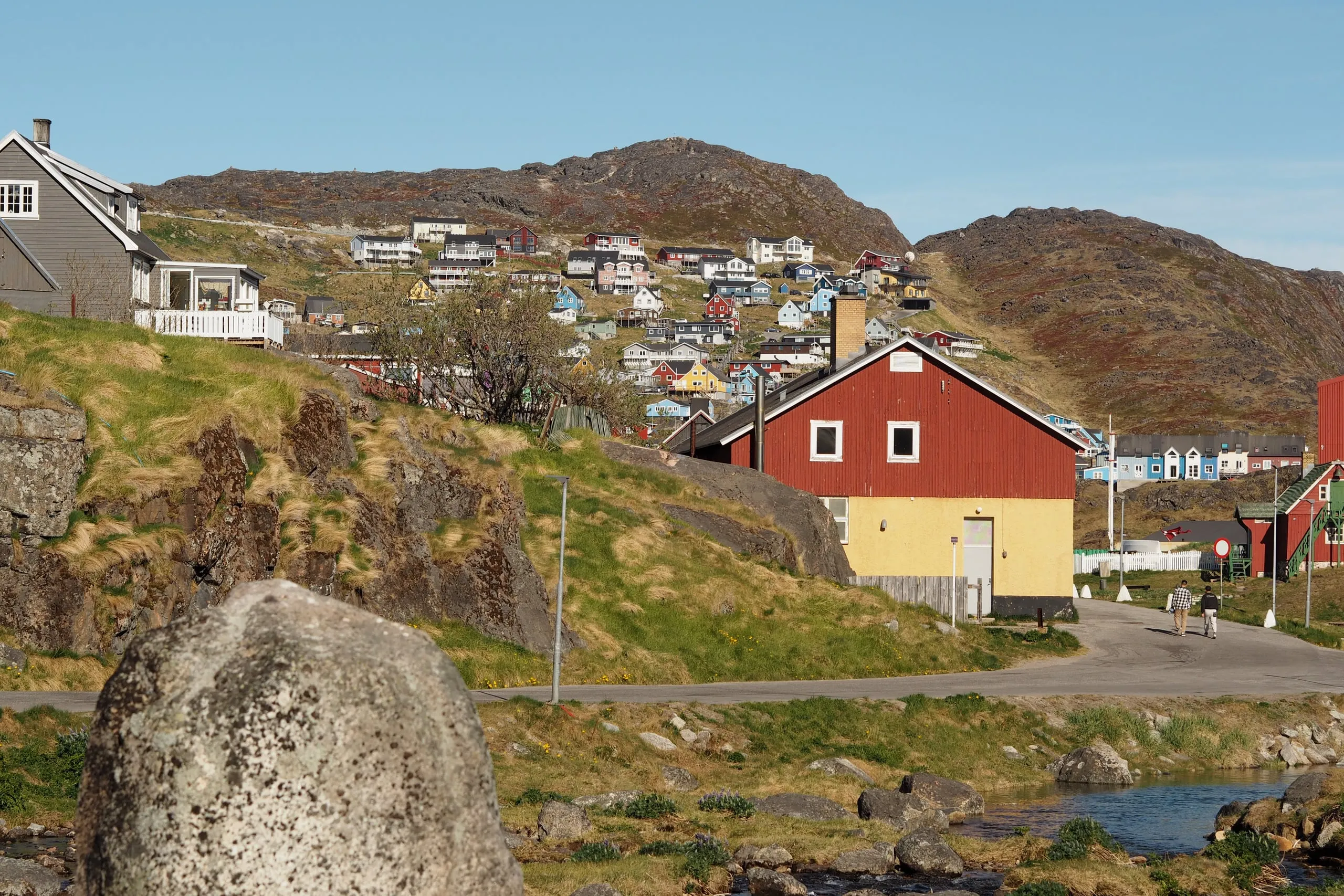 A red and yellow house beside a stream, with houses dotted over the rocky hills behind.
