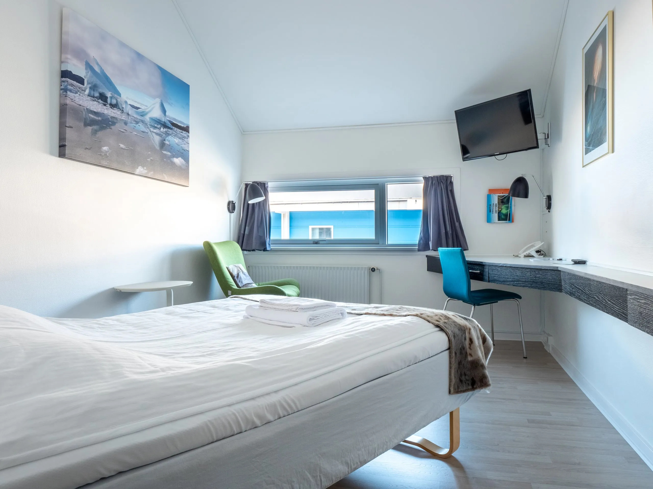 A single room with the bed under a fur throw, a long desk beneath the TV, and an iceberg photograph on the wall.