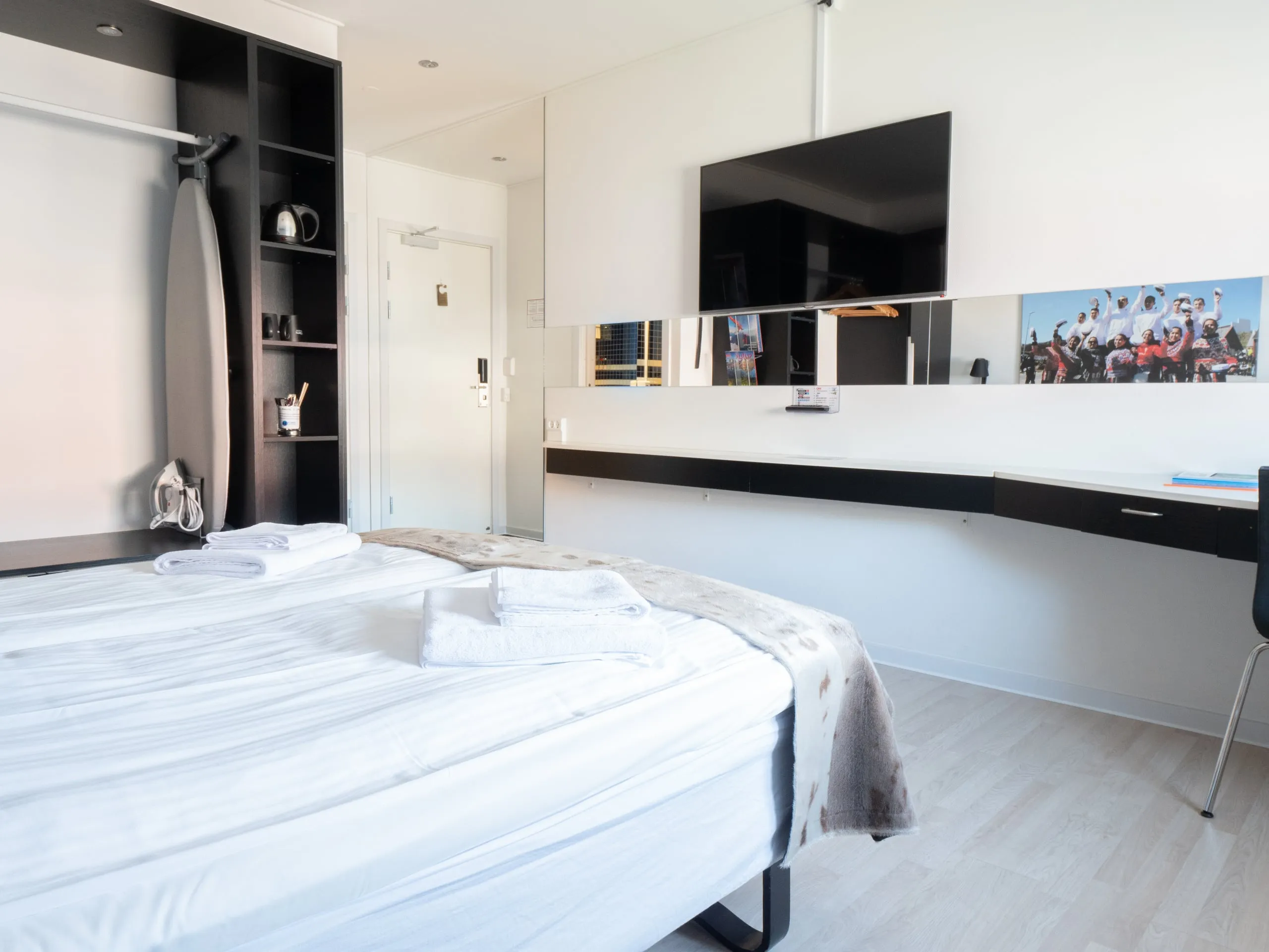 A double room with the bed in front of a long desk, a TV on the wall and an open wardrobe.