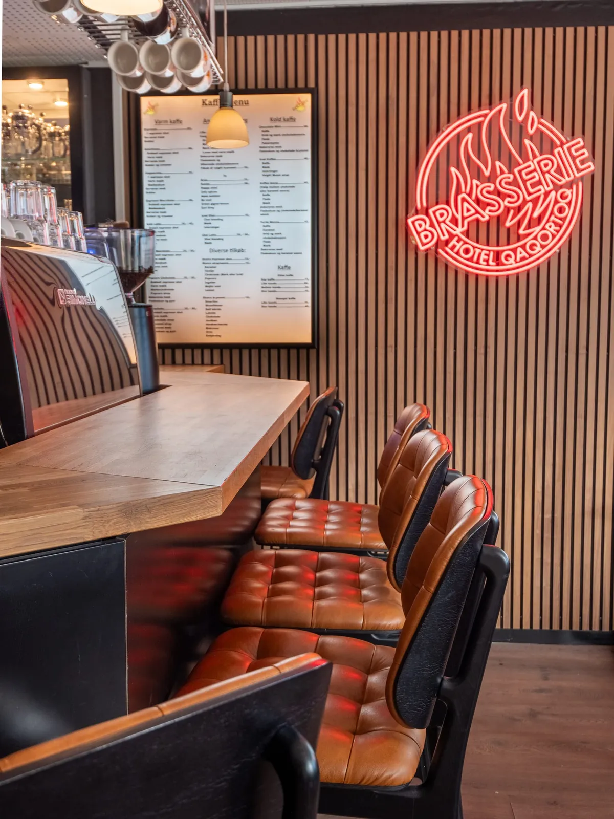 The coffee bar in the brasserie: a wooden counter with leather stools, and the red neon sign of Brasserie Hotel Qaqortoq.