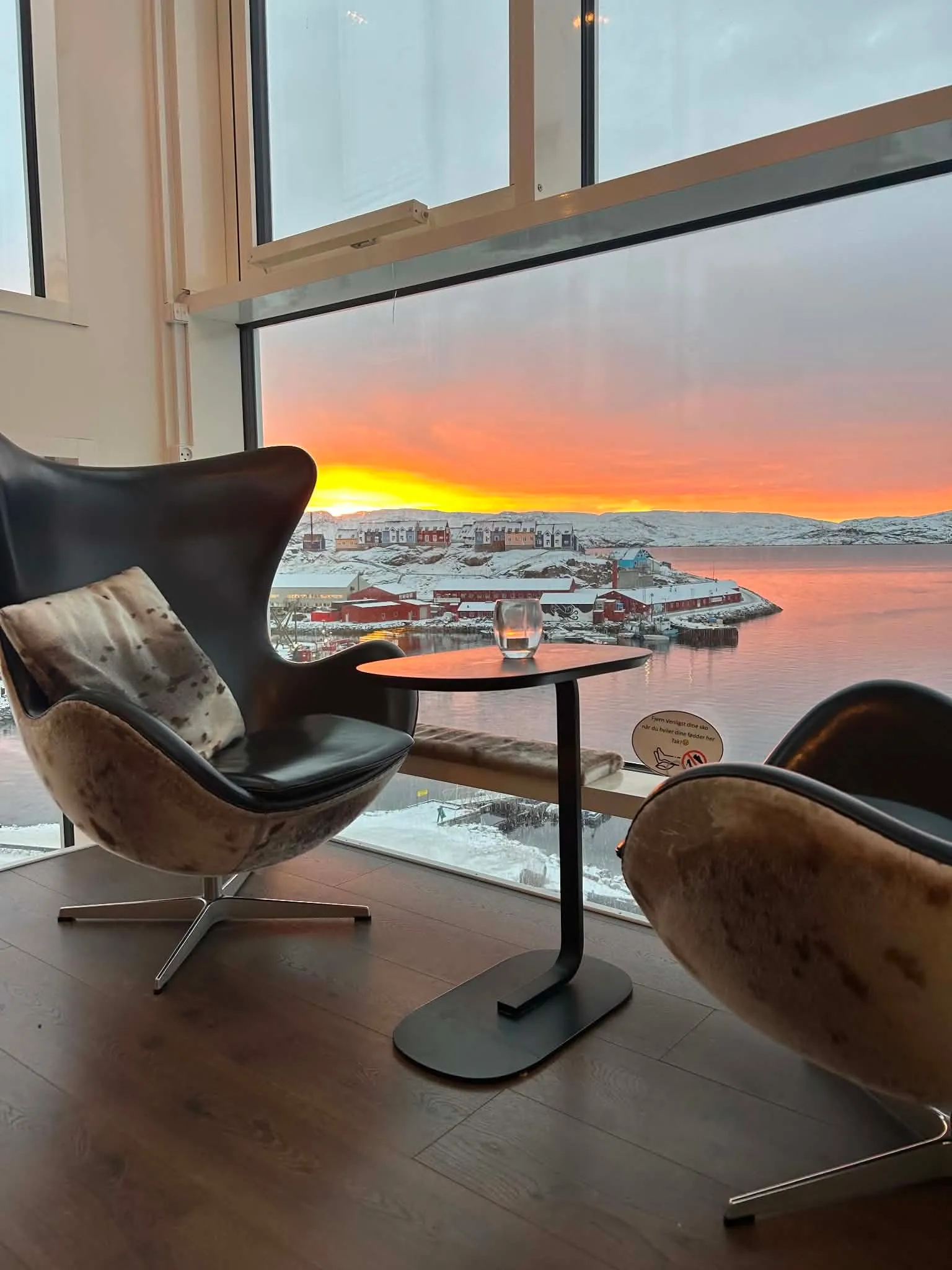 Two egg chairs and a small table by a window, the sky orange over the snow-covered harbour.
