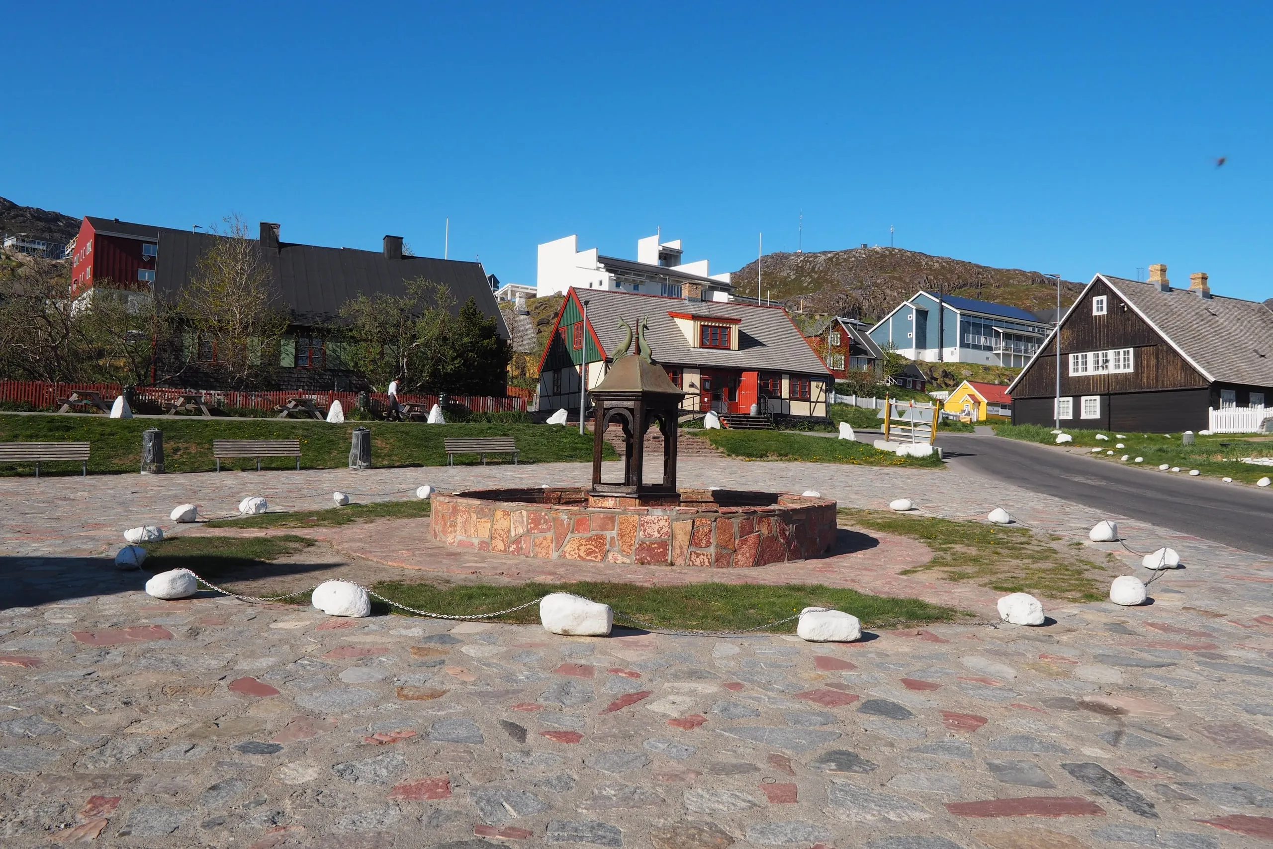 The town square and its old fountain, ringed by white stones, in front of timber houses.