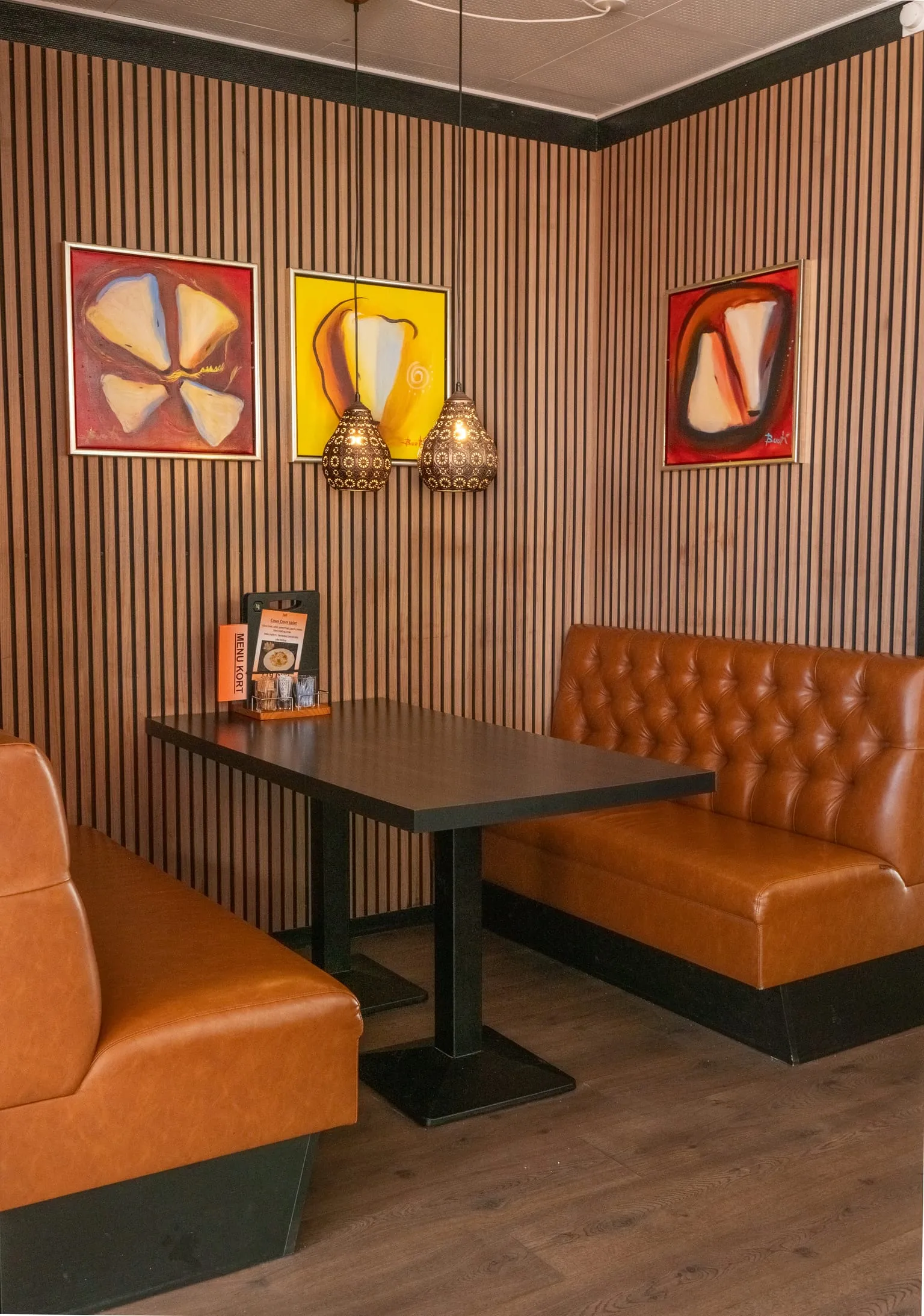 A corner booth in tan leather against a slatted wooden wall hung with three bright paintings.