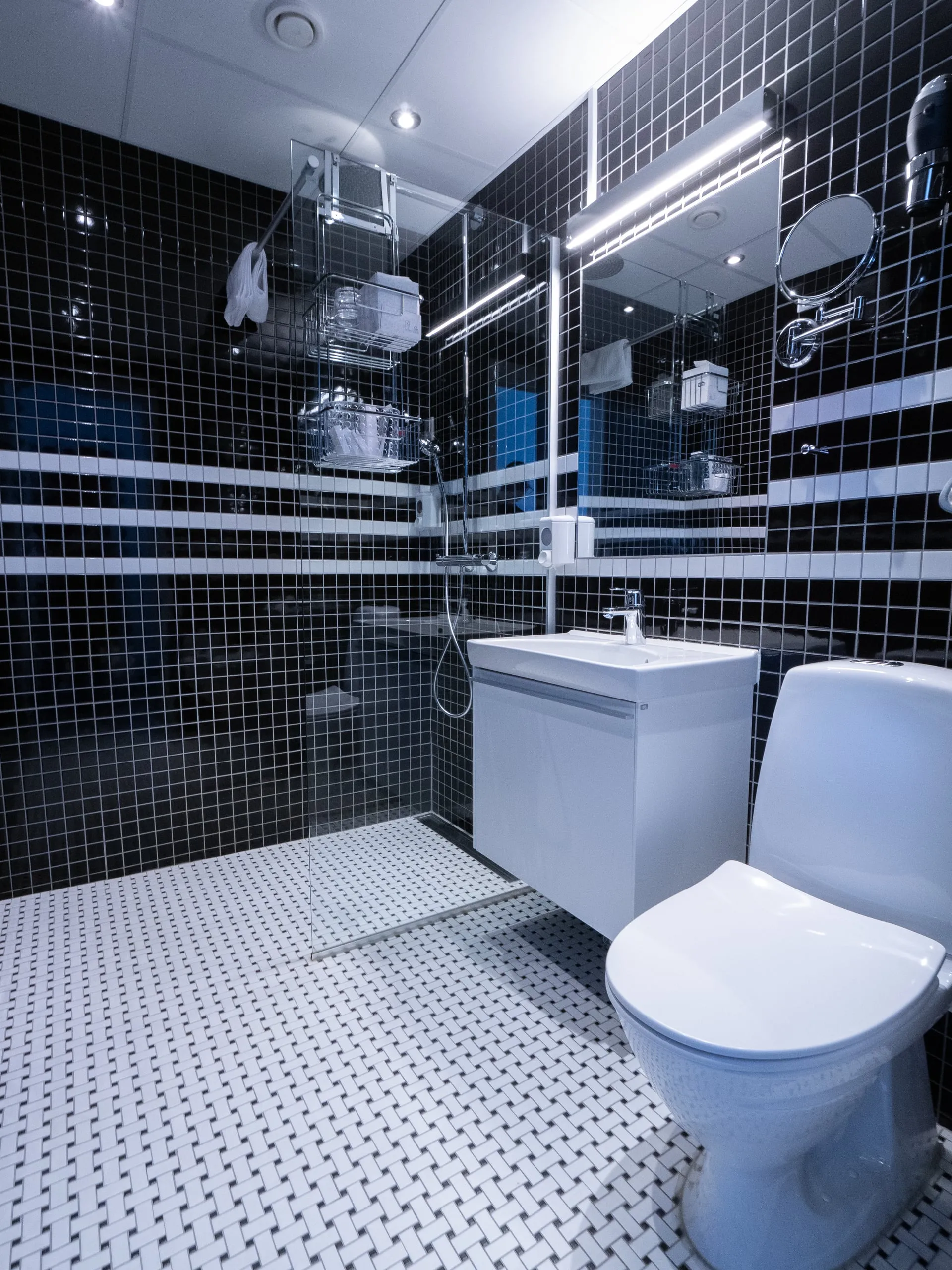 The bathroom of a double room, with a basin, a toilet and a shower against black and white tiles.