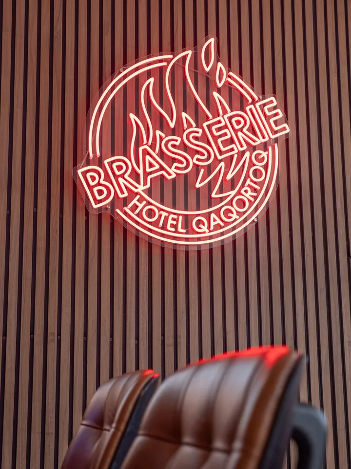 The red neon sign of Brasserie Hotel Qaqortoq on a slatted wooden wall, above leather bar stools.