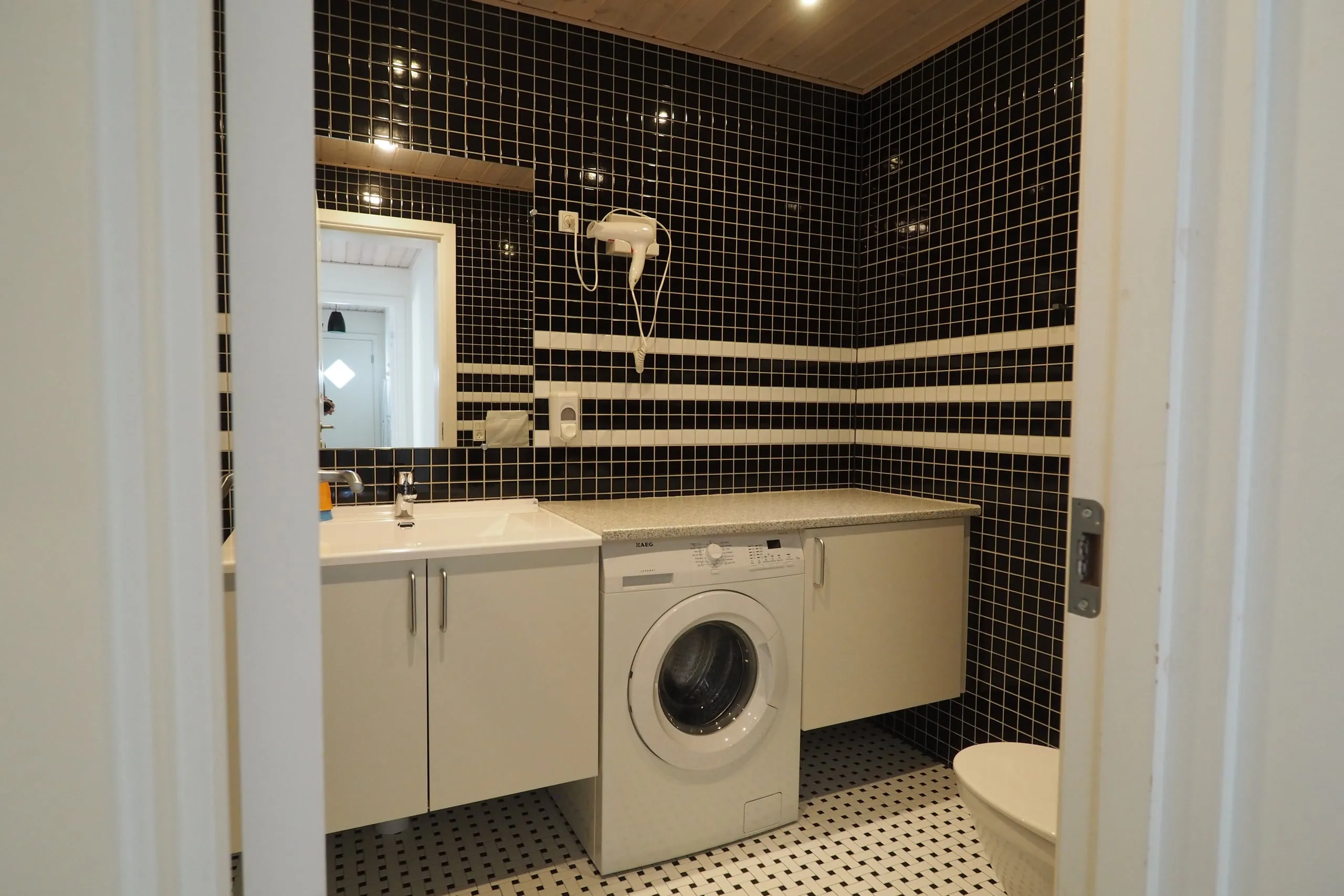 An apartment bathroom seen from the door, with a basin, a washing machine and black and white tiles.
