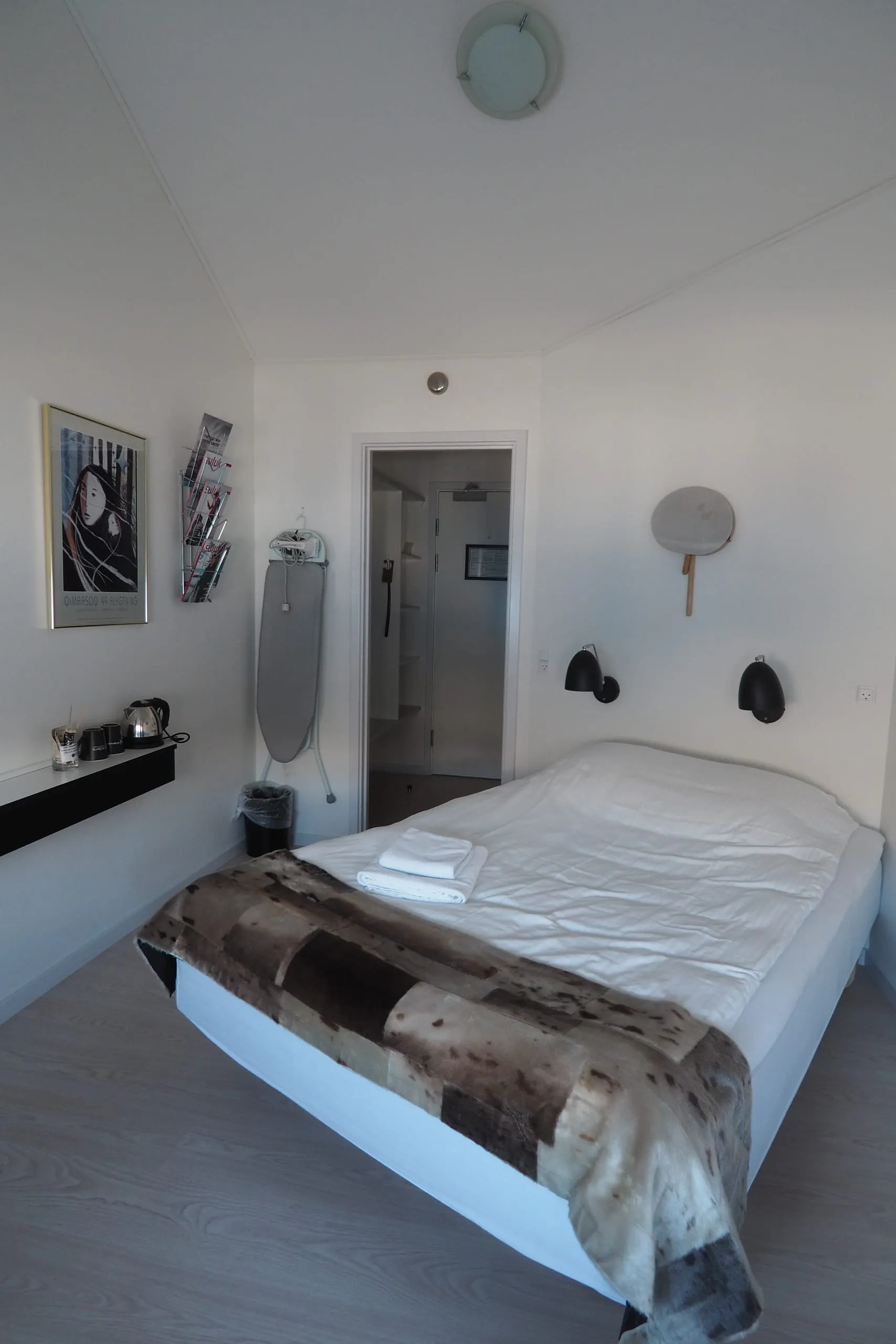 A single room seen from the window: the full bed under a fur throw, reading lamps above it, and the door out to the hallway.