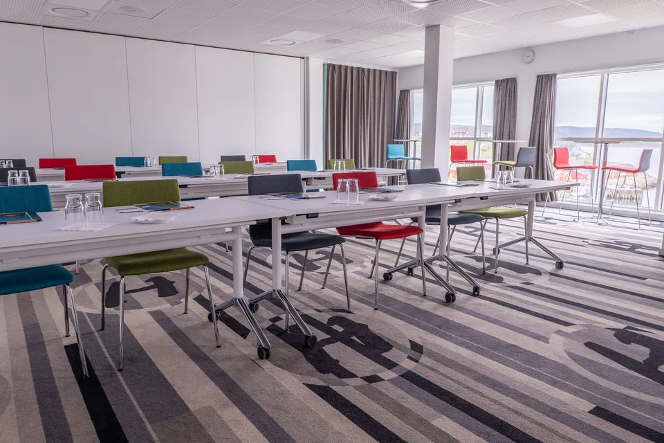 The conference venue set with rows of tables and colourful chairs, its tall windows looking out over Qaqortoq.